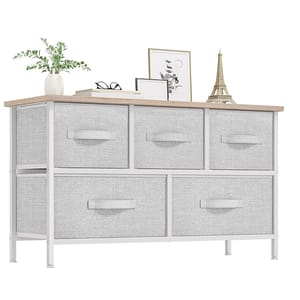 A 5-drawer dresser with a light gray fabric design and a wooden top, featuring three smaller top drawers and two larger bottom drawers. The dresser is accessorized with a plant, glasses, books, framed art, and an Eiffel Tower model.