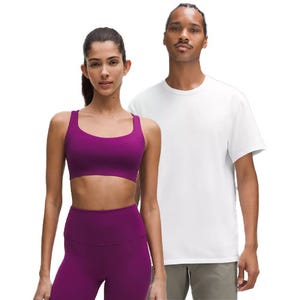 A woman in a purple athletic outfit, consisting of a sports bra and leggings, stands next to a man wearing a white T-shirt and beige pants.
