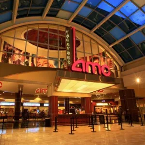 AMC Theatres entrance with bright signage, a large glass atrium, and poster displays. Queue barriers are set up in front of the ticket area.