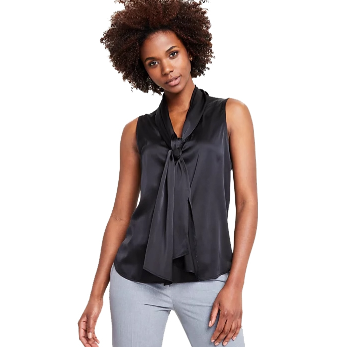A woman is wearing a sleek sleeveless black blouse with a prominent bow at the neckline, paired with light gray pants.