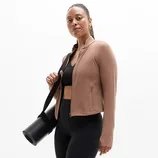 A person wearing a fitted brown jacket over a black sports bra and black leggings, carrying a black yoga mat with a shoulder strap.