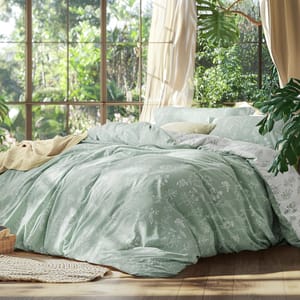 A cozy bedroom features a bed with light green floral-patterned bedding and a cream throw. Large windows reveal lush greenery outside, enhancing the serene atmosphere.