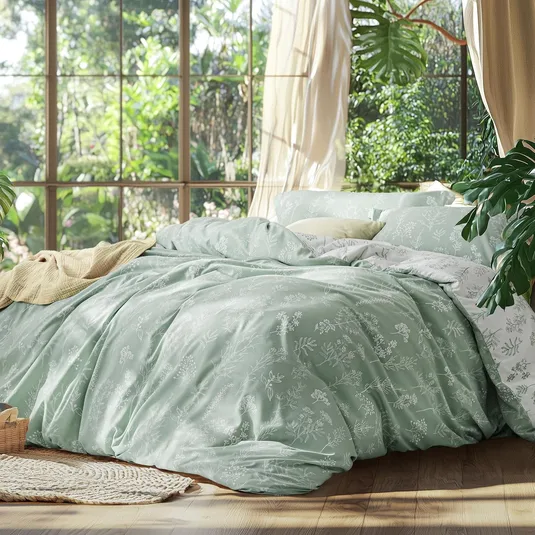 A cozy bedroom features a bed with light green floral-patterned bedding and a cream throw. Large windows reveal lush greenery outside, enhancing the serene atmosphere.