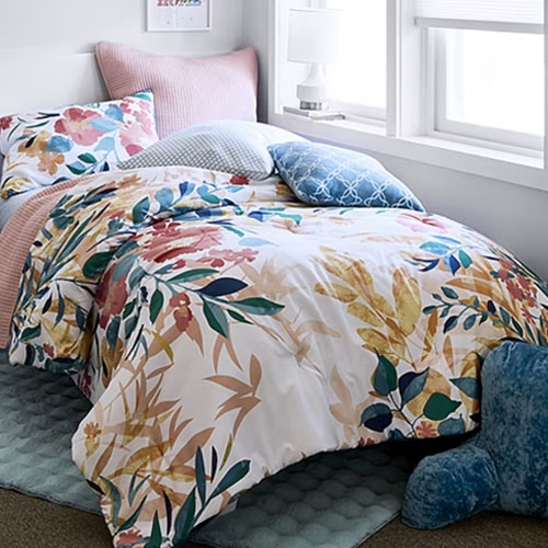 Floral-patterned bedding set on a bed with multiple pillows, including pink striped and blue textured designs, in a bright room with a window.