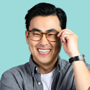 A person wearing a light-colored shirt, brown-rimmed glasses, and a black wristwatch against a blue background.