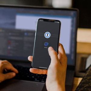 A smartphone screen displays the 1Password logo with text indicating a 25% discount for the first year, against the backdrop of a laptop being used by a person.