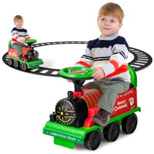 A child is riding a green and red Christmas-themed toy train on a circular track. The train features a steering wheel and festive decorations with \“Merry Christmas\“ text and Santa graphics.