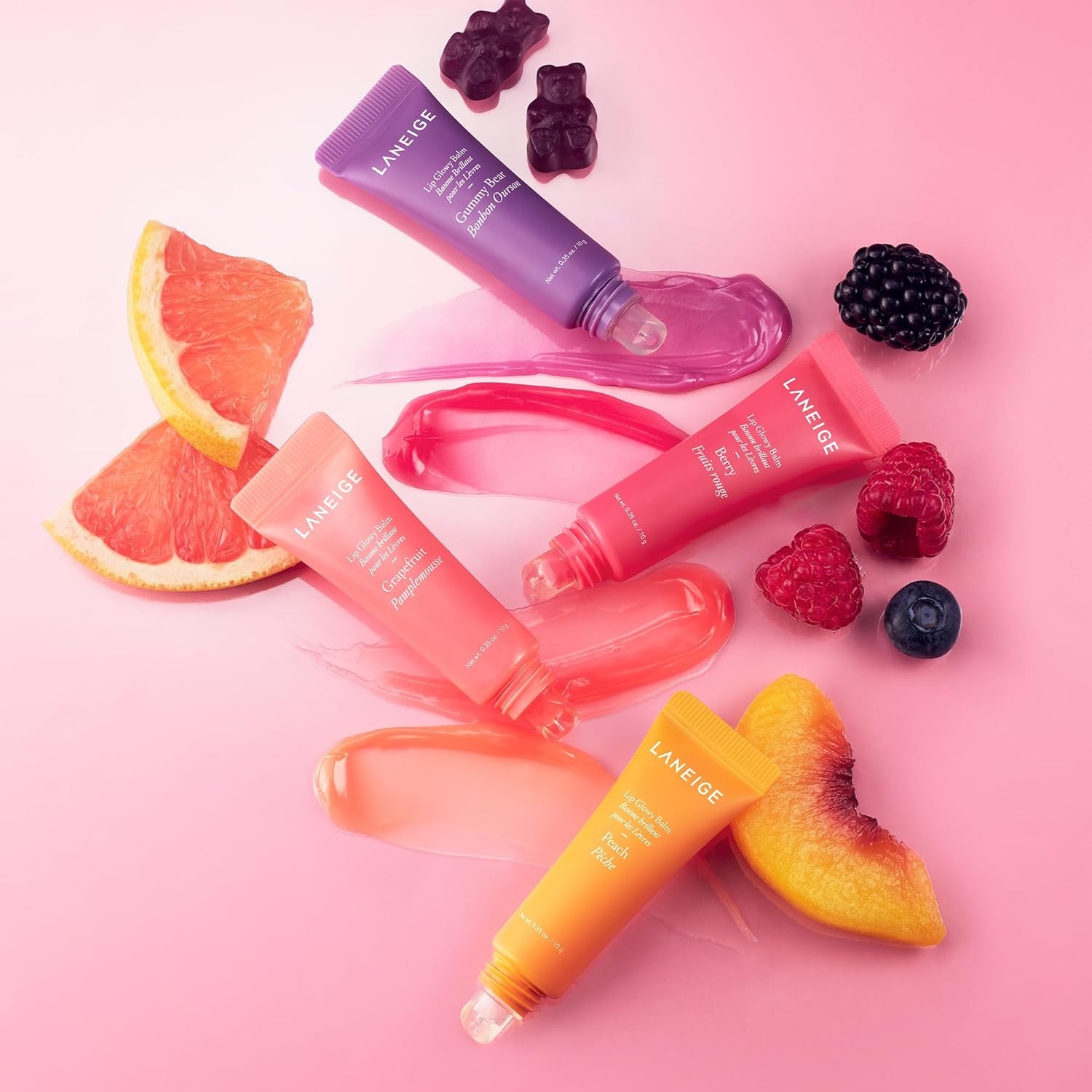 Three colorful tubes of lip gloss are displayed amidst fruit slices and berries on a pink background.