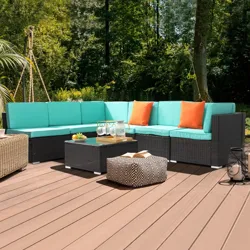 Outdoor patio set with a wicker sectional sofa featuring turquoise cushions and orange pillows, accompanied by a glass-top coffee table and patterned pouf on a wooden deck surrounded by greenery.