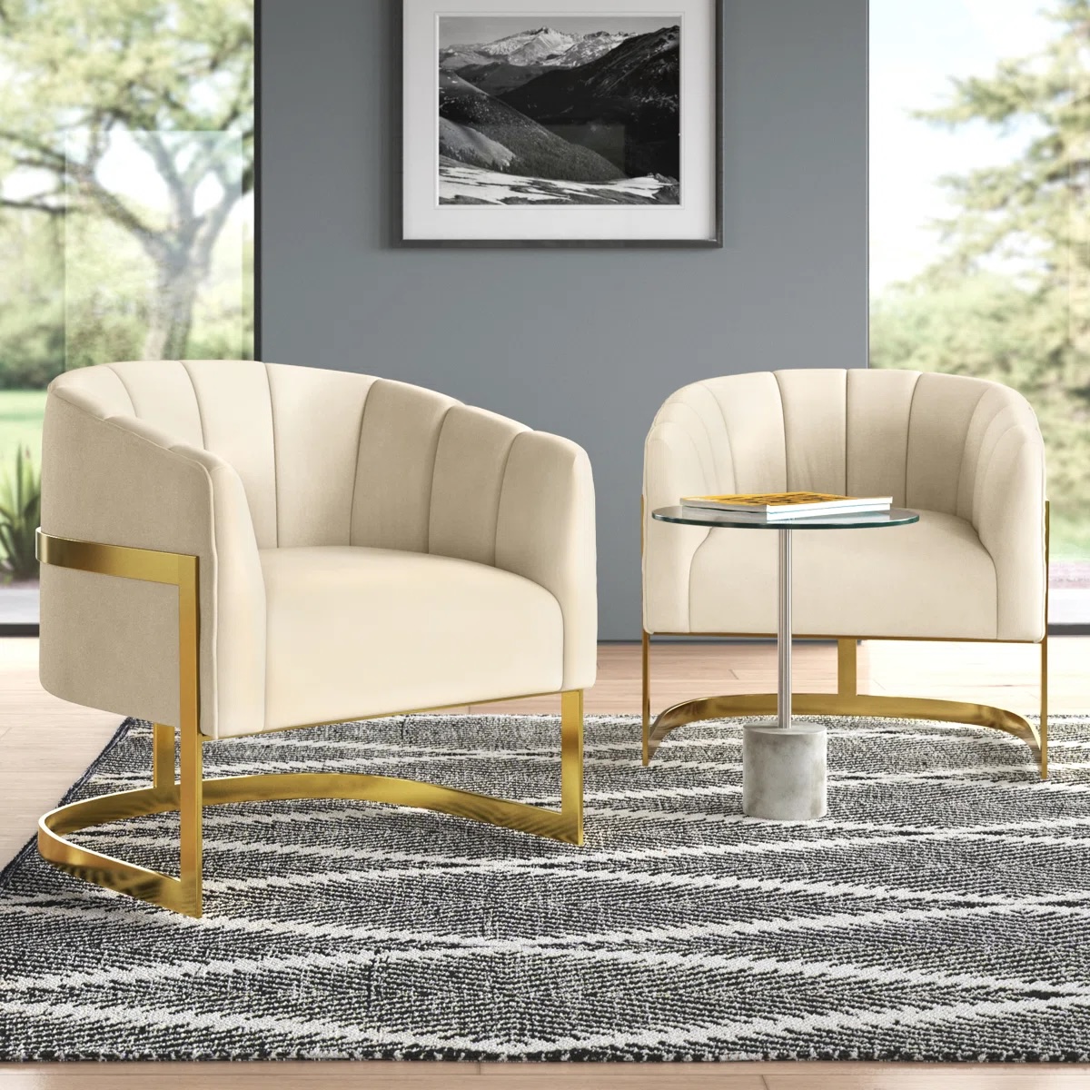 Two cream-colored, modern barrel chairs with gold metal accents are paired with a small round glass side table featuring a marble base. They are placed on a patterned gray rug in a well-lit room with large windows and a landscape photograph on the wall.