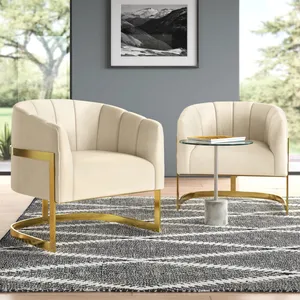 Two cream-colored, modern barrel chairs with gold metal accents are paired with a small round glass side table featuring a marble base. They are placed on a patterned gray rug in a well-lit room with large windows and a landscape photograph on the wall.