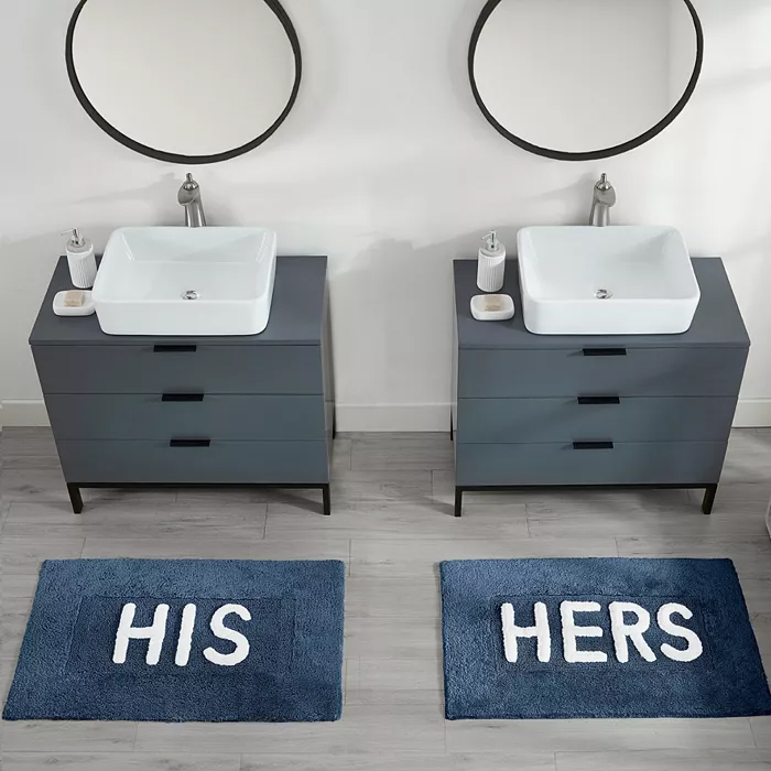Double bathroom vanity set featuring two circular mirrors, white rectangular sinks, and gray cabinets, with blue rugs labeled \“His\“ and \“Hers\“ placed on a light gray floor.