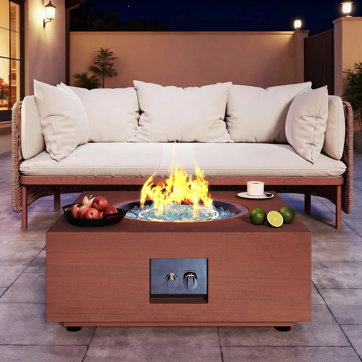 A patio with a modern fire pit table featuring a flame surrounded by blue glass, accompanied by a cup and limes. Nearby, a cushioned outdoor sofa and a bowl of apples sit on paved flooring.