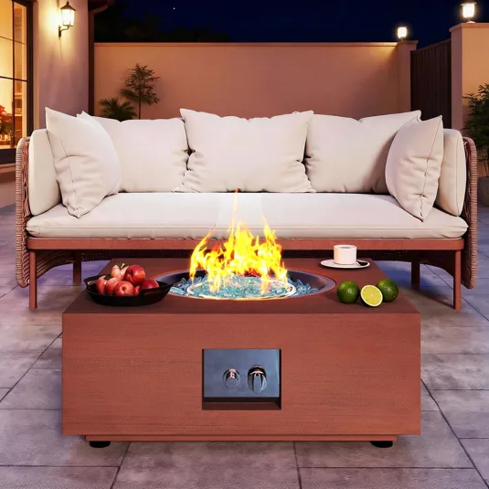 A patio with a modern fire pit table featuring a flame surrounded by blue glass, accompanied by a cup and limes. Nearby, a cushioned outdoor sofa and a bowl of apples sit on paved flooring.