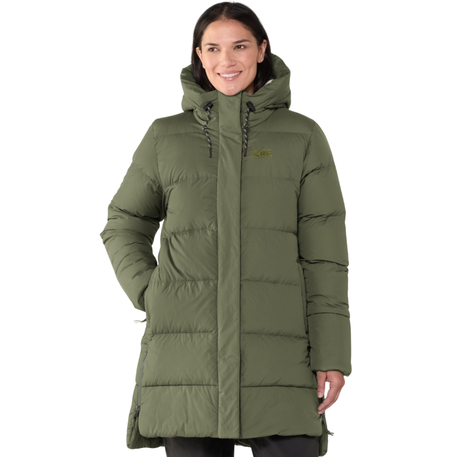 A woman is wearing an olive green, hooded, puffer coat with quilted stitching and hand pockets. The coat features a logo on the upper chest.