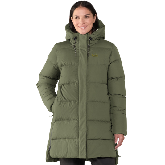 A woman is wearing an olive green, hooded, puffer coat with quilted stitching and hand pockets. The coat features a logo on the upper chest.