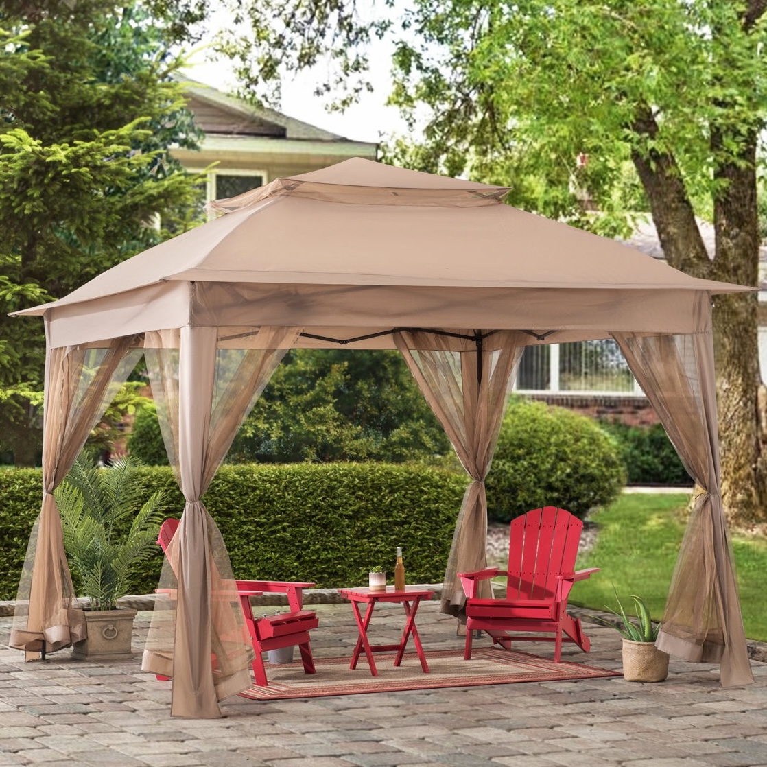 A beige gazebo with mesh netting encloses two red Adirondack chairs, a small red table, a rug, and potted plants, set on a paved patio.