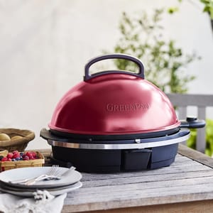 A red GreenPan electric grill is on a wooden table, surrounded by tableware and a basket of berries.