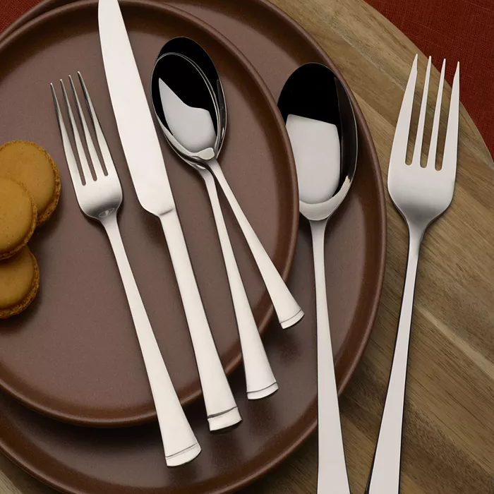 A set of silver flatware including a knife, fork, spoon, and two smaller spoons is displayed on brown ceramic plates, accompanied by a few small cookies on a wooden surface.