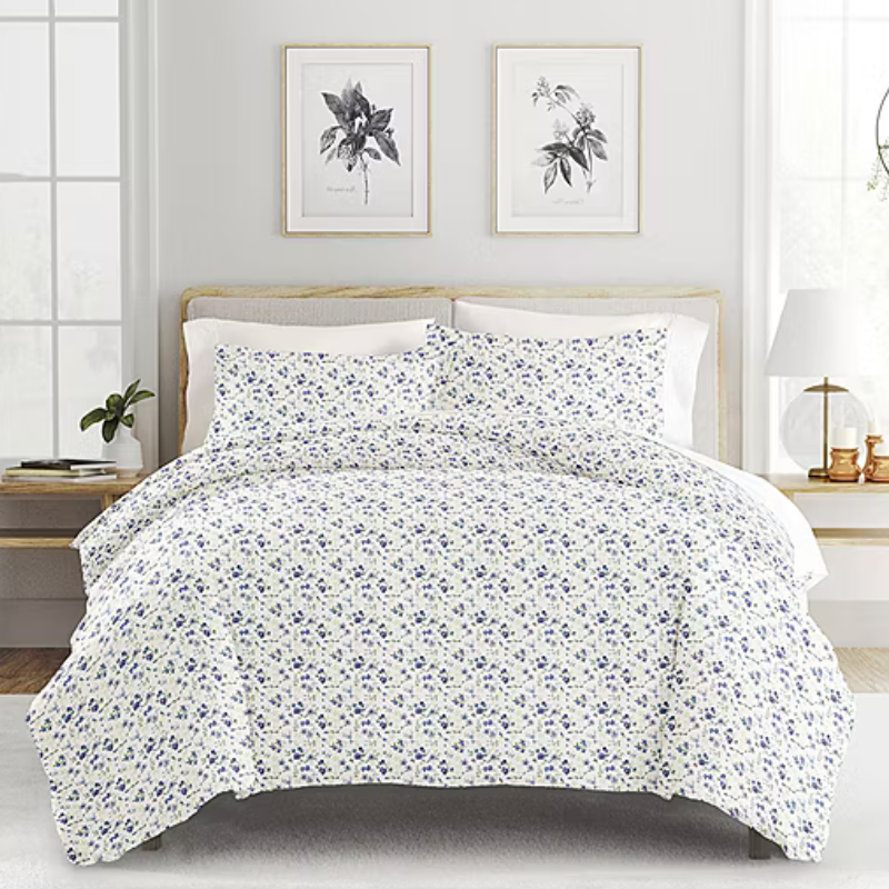 A bed features a floral-patterned comforter set with matching pillowcases in a light and airy bedroom. The room includes framed botanical prints, a wooden nightstand, potted plant, table lamp, and large windows.