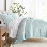 A bed features a reversible comforter set with a light blue and gray design, accompanied by matching pillows. The bedding is complemented by sheer curtains and a wooden nightstand with a plant, creating a tranquil and cozy bedroom ambiance.