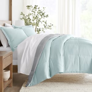 A bed features a reversible comforter set with a light blue and gray design, accompanied by matching pillows. The bedding is complemented by sheer curtains and a wooden nightstand with a plant, creating a tranquil and cozy bedroom ambiance.
