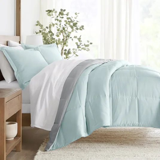 A bed features a reversible comforter set with a light blue and gray design, accompanied by matching pillows. The bedding is complemented by sheer curtains and a wooden nightstand with a plant, creating a tranquil and cozy bedroom ambiance.