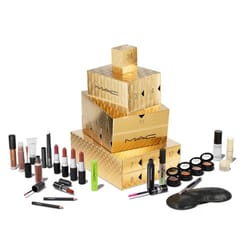 24-piece MAC Advent Calendar features gold stacked boxes with numbered compartments, containing a variety of lipsticks, lip glosses, eyeshadows, mascaras, eyeliners, face masks, and nail polishes in assorted colors and finishes.