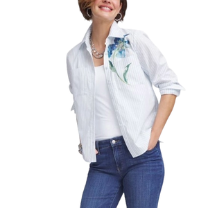 A woman wears a casual outfit with a white tank top under a pinstriped shirt featuring a floral design on the shoulder, paired with blue jeans.