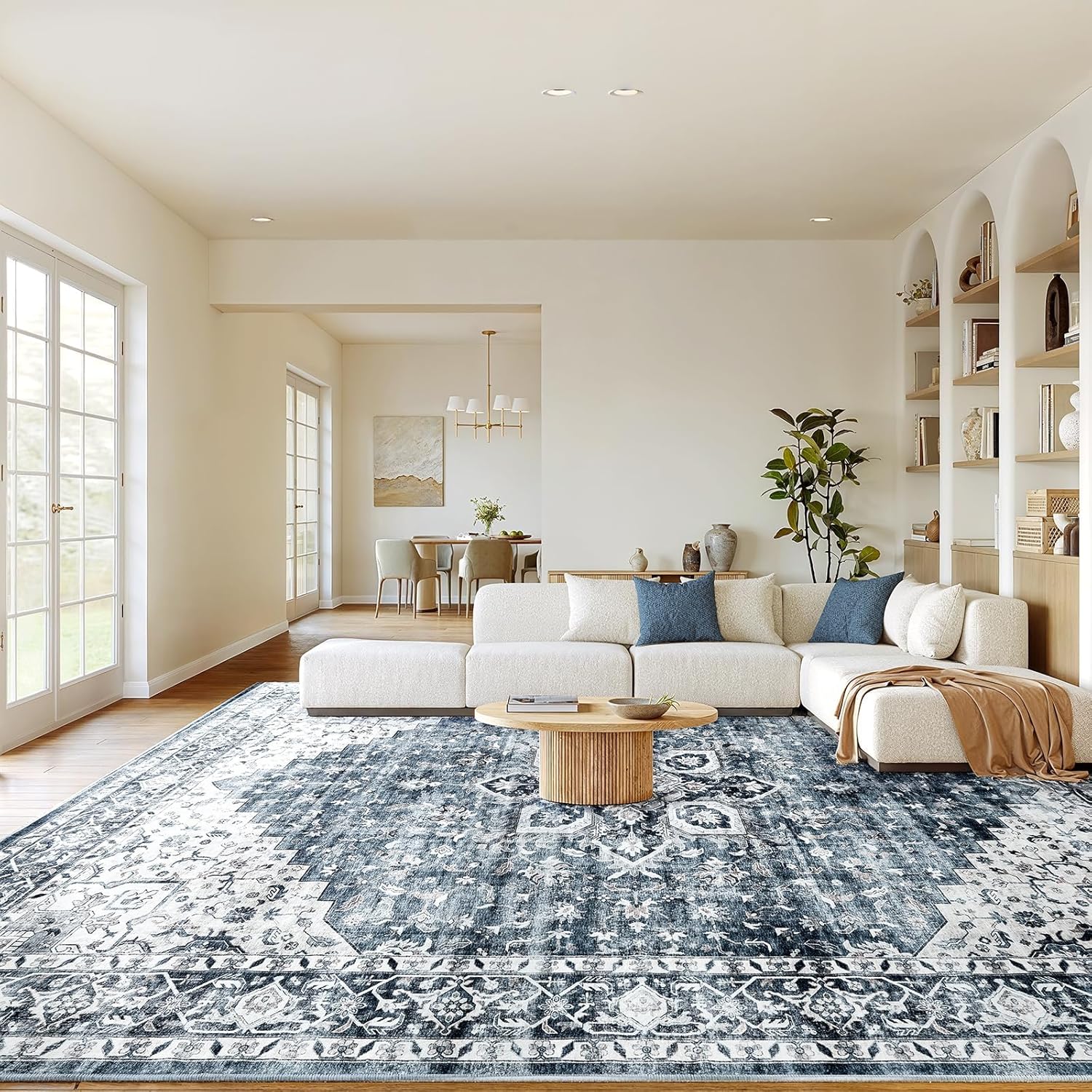 A spacious living room features a large blue and white patterned rug, a cream sectional sofa with blue pillows, a round wooden coffee table, and built-in shelves. A plant adds greenery to the neutral-toned decor.