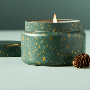 A lit green tin candle with decorative yellow and gray patterns is placed on a surface alongside a small pine cone.
