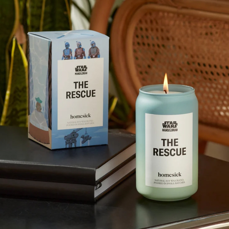 A lit candle with \“THE RESCUE\“ label beside its boxed packaging, both branded with \“Star Wars: The Mandalorian.\“
