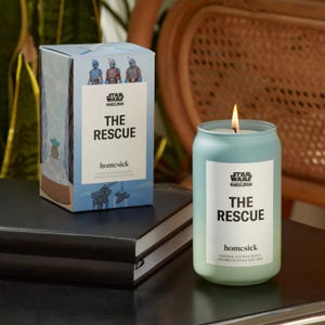 A lit candle with \“THE RESCUE\“ label beside its boxed packaging, both branded with \“Star Wars: The Mandalorian.\“