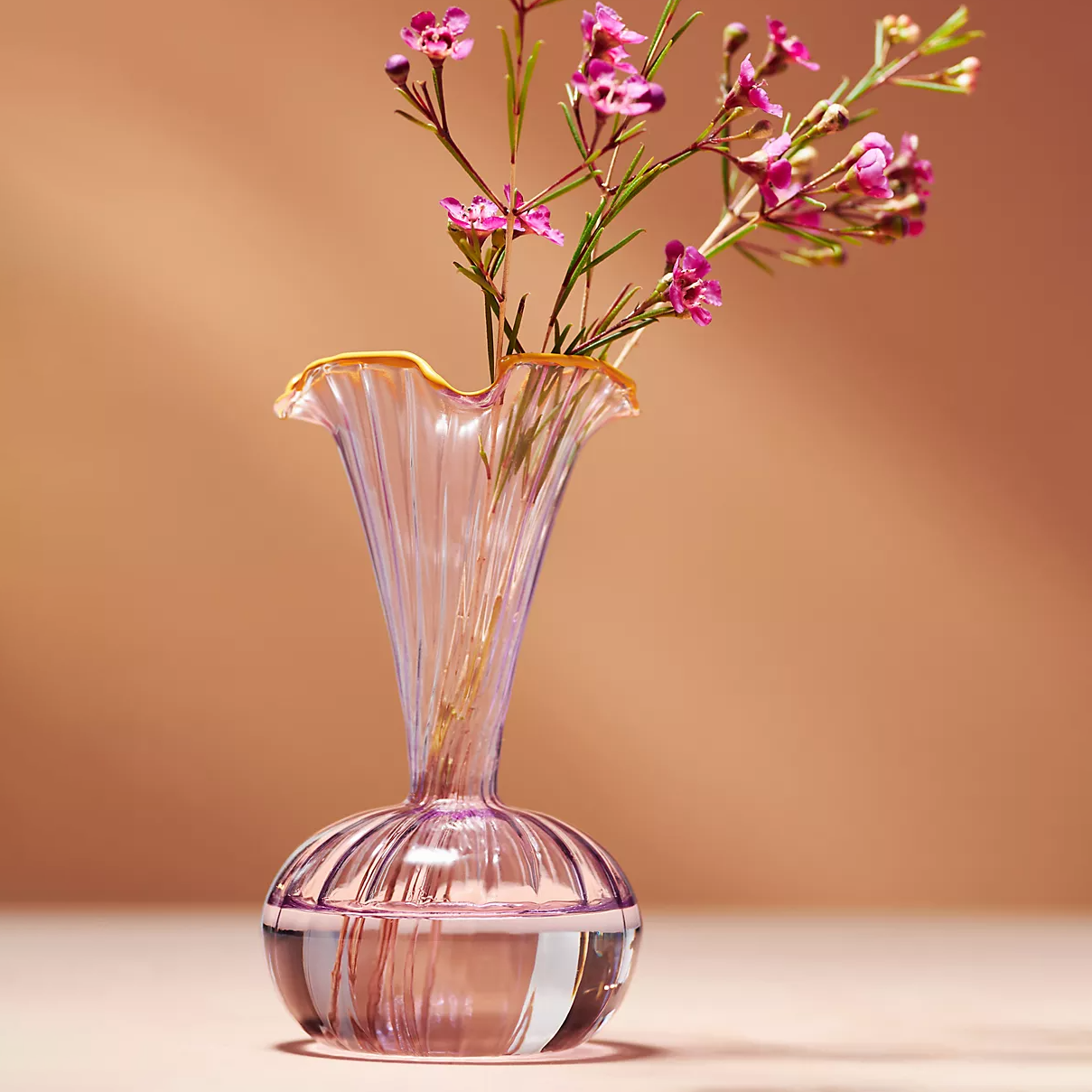 A translucent pink glass vase with an elegant flared rim and ribbed detailing holds a spray of pink flowers.