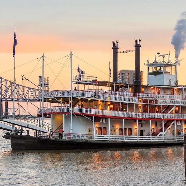 A paddle steamer with multiple decks docked by the river at dusk.