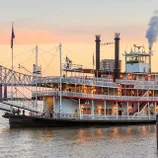 A paddle steamer with multiple decks docked by the river at dusk.