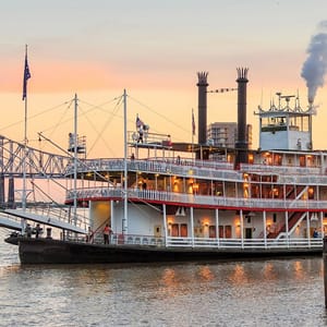 A paddle steamer with multiple decks docked by the river at dusk.