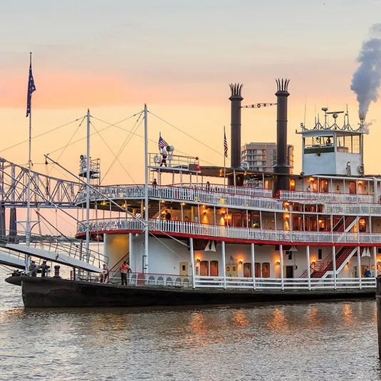 A paddle steamer with multiple decks docked by the river at dusk.
