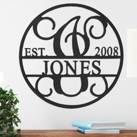 A round, black metal wall sign with decorative swirls features the words \“EST 2008\“ and \“JONES\“ with a large ornamental letter \“J\“ in the center, alongside a small stack of books on a surface.