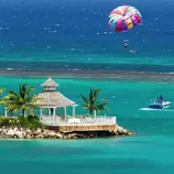 A tropical beach scene with clear blue waters, a white gazebo, palm trees, and parasailing in the background.