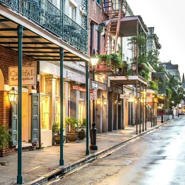 A charming street scene showcases storefronts with classic Southern architecture, featuring ornamental iron balconies and inviting shop signs. Softly lit cobblestones reflect a peaceful, early evening ambiance, enhanced by warm streetlights.