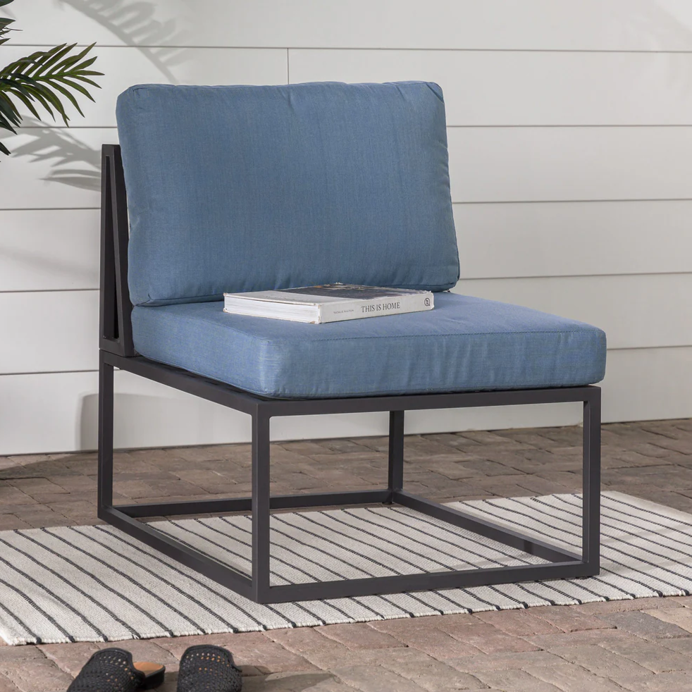 A blue cushioned chair with a dark metal frame sits on a striped rug on a brick patio. There's a book titled \“This is Home\“ on the seat, and a pair of black woven slippers nearby.
