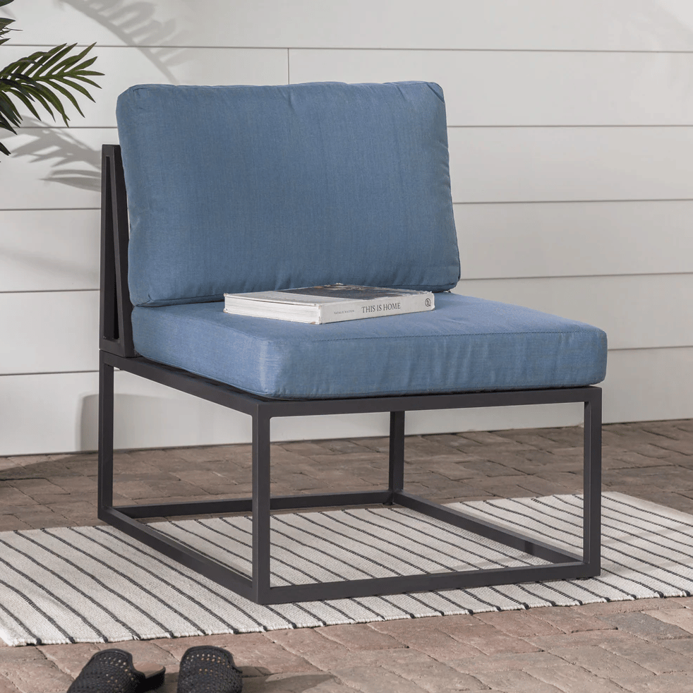 A blue cushioned chair with a dark metal frame sits on a striped rug on a brick patio. There's a book titled \“This is Home\“ on the seat, and a pair of black woven slippers nearby.