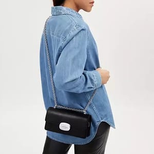 A person wearing a light blue denim shirt and black trousers with a small black shoulder bag featuring a chain strap.