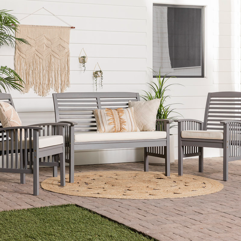 A patio set with a bench, two chairs, and decorative pillows on a round rug, against a white house with hanging plants and a woven wall hanging.