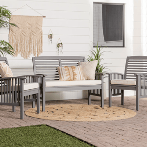 A patio set with a bench, two chairs, and decorative pillows on a round rug, against a white house with hanging plants and a woven wall hanging.