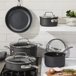A cookware set featuring a nonstick frying pan, two saucepans with lids, and a stockpot with a lid, displayed on a kitchen stove and shelf with a cutting board and herbs nearby.