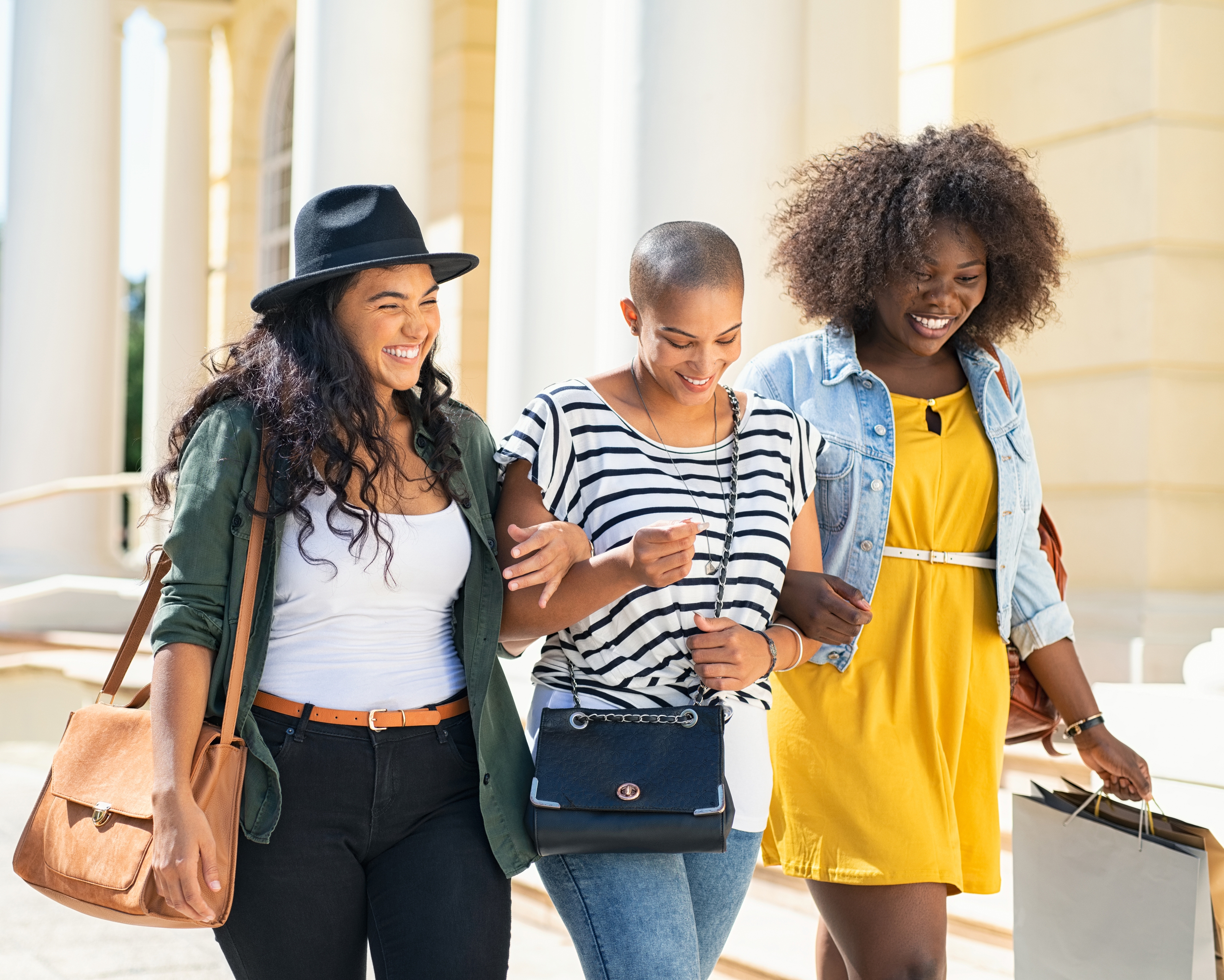 Three women are walking together, two carrying shoulder bags and one holding shopping bags.