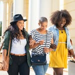 Three women are walking together, two carrying shoulder bags and one holding shopping bags.
