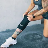 A person wearing a knee sleeve with the logo \“X\“ while adjusting it over a tattooed leg. They are wearing white athletic shoes, a smartwatch, and a sleeveless top.